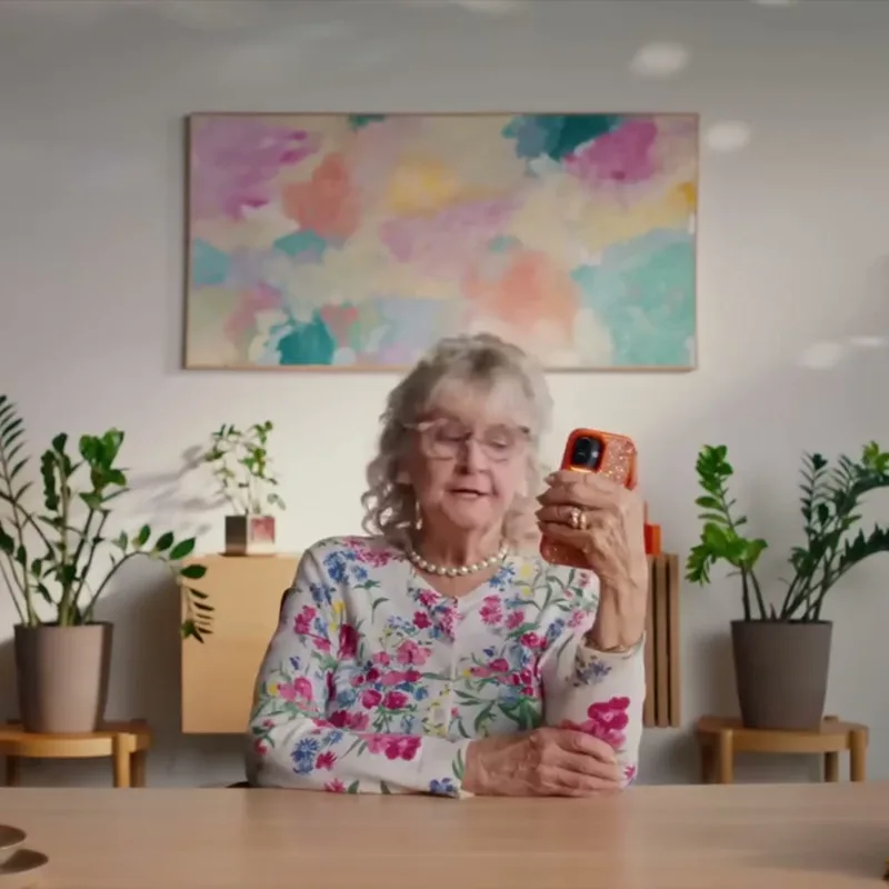 An older woman in a floral cardigan sitting at a wooden table, holding up an orange phone and talking to it