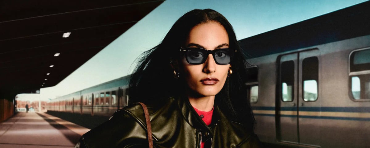 A woman on a train platform wearing Ray-Ban Meta Audio, black rectangular frames with smoked blue lenses and no camera