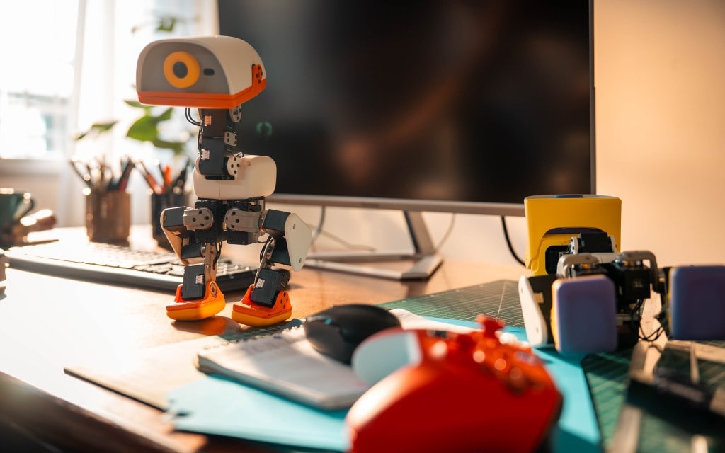 A single white and orange Microduck robot standing on a desk beside a keyboard, monitor, game controller and spare parts