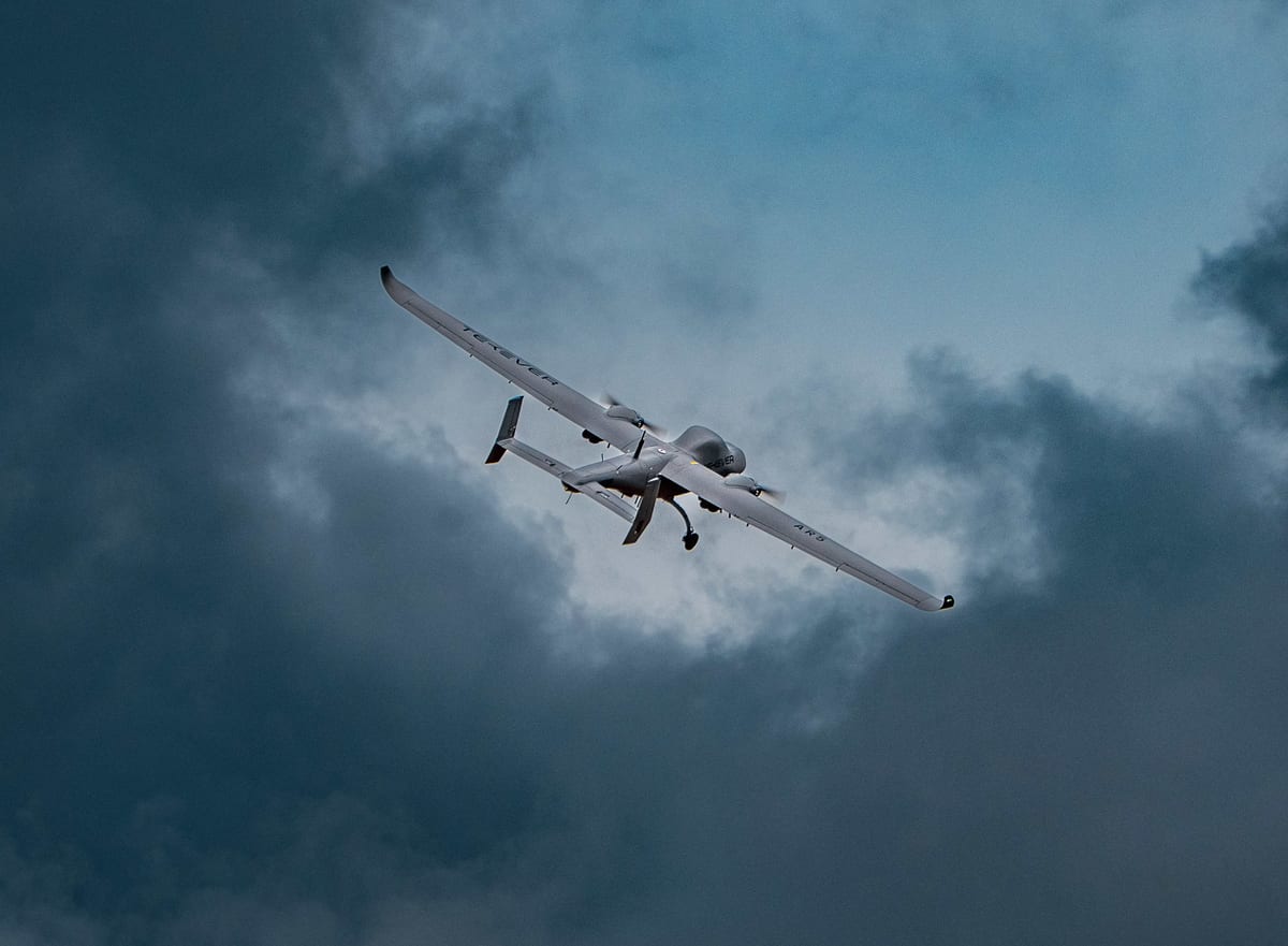 A grey Tekever AR5 fixed-wing drone banks through a break in dark storm clouds