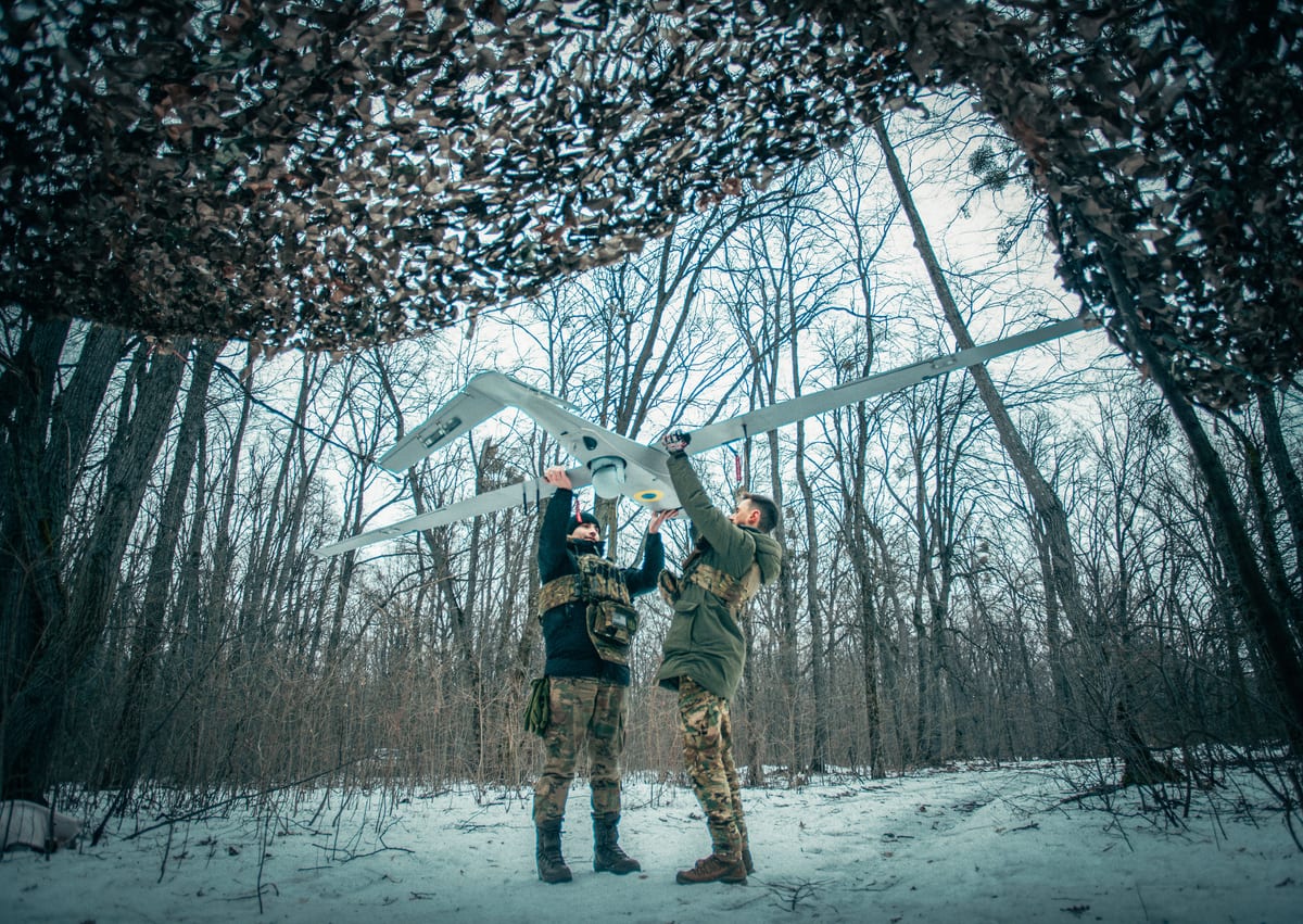 Two operators in camouflage stand in a snowy forest under camouflage netting, holding a white Tekever fixed-wing drone overhead